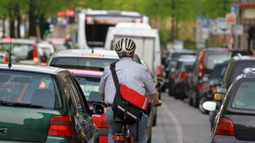 Ein Radfahrer mit Helm und roter Umhängetasche fährt ohne Fahrradweg in dichten Stadtverkehr